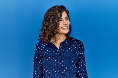 Kıvırcık saçlı, mavi arka planda günlük kıyafetler giyen, yüzünde gülümsemeyle yan yana bakan esmer bir kadın, doğal bir ifade. Kendinden emin gülen. 