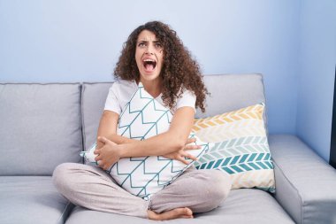Hispanic woman with curly hair sitting on the sofa at home angry and mad screaming frustrated and furious, shouting with anger looking up. 