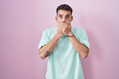 Pembe arka planda duran yakışıklı İspanyol adam yanlışlıkla elleriyle ağzını kapatırken şok oldu. gizli kavram. 