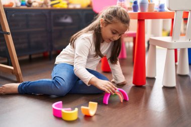 Adorable hispanic girl playing with toys sitting on floor at kindergarten