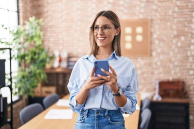 Genç, güzel İspanyol iş kadını ofisteki akıllı telefonu kullanarak gülümsüyor.