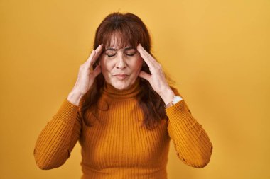 Middle age hispanic woman standing over yellow background with hand on head, headache because stress. suffering migraine. 