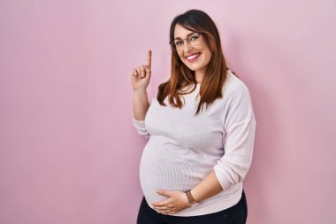 Pregnant woman standing over pink background showing and pointing up with finger number one while smiling confident and happy. 