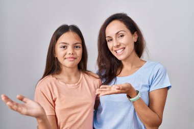 Young mother and daughter standing over white background pointing aside with hands open palms showing copy space, presenting advertisement smiling excited happy 
