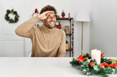 Young handsome man with beard sitting on the table by christmas decoration very happy and smiling looking far away with hand over head. searching concept. 