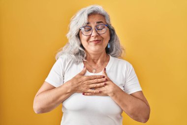 Sarı arka planda gri saçlı, elleri göğsünde, gözleri kapalı ve yüzünde minnet ifadesi olan orta yaşlı bir kadın. sağlık kavramı. 