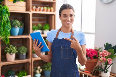 Brunette young woman working at florist shop holding tablet smiling happy pointing with hand and finger 