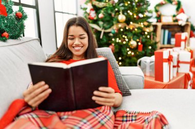 Genç Latin kadın kitap okuyor. Yılbaşı ağacının yanında oturuyor.