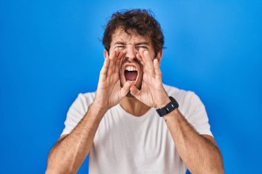 Mavi arka planda dikilen Latin asıllı genç adam elleri ağzında kızgın bir şekilde bağırıyor. 