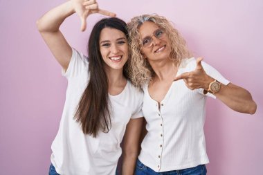 Anne ve kızı pembe arka planda bir arada durup gülümsüyor ve mutlu yüzlü eller ve parmaklarla çerçeve yapıyorlar. yaratıcılık ve fotoğrafçılık kavramı. 