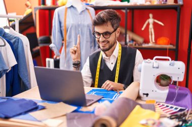 Dövmeli genç İspanyol adam terzi tasarımcısı terzi odasında dizüstü bilgisayar kullanıyor. Bir fikirle gülümsüyor ya da soru işareti yapıyor. 