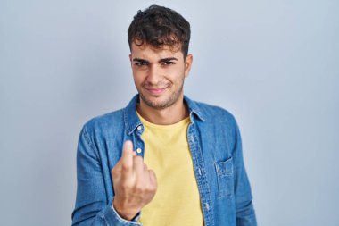 Young hispanic man standing over blue background beckoning come here gesture with hand inviting welcoming happy and smiling 