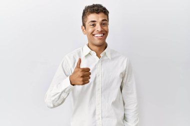 Young handsome hispanic man standing over isolated background doing happy thumbs up gesture with hand. approving expression looking at the camera showing success. 