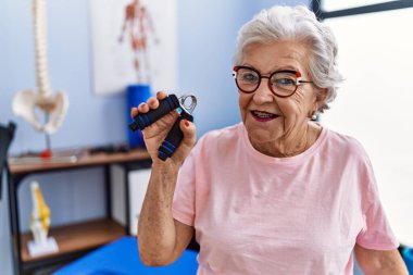 Yaşlı gri saçlı kadın fizyoterapist terapi kliniğinde el ele tutuşarak rehabilitasyon seansı yapıyor.