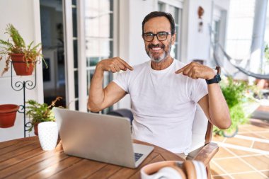 Orta yaşlı bir adam bilgisayarı evde kullanıyor. Yüzünde gülümsemeyle kendinden emin görünüyor. Kendini gururlu ve mutlu gösteriyor.. 