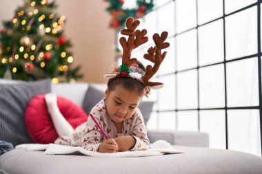 Adorable hispanic girl drawing on notebook lying on sofa by christmas tree at home