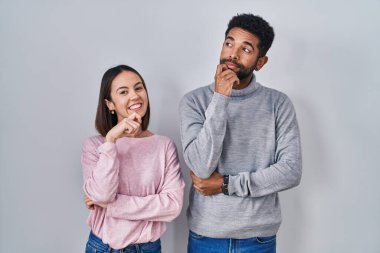 Young hispanic couple standing together with hand on chin thinking about question, pensive expression. smiling and thoughtful face. doubt concept. 