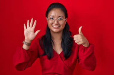 Asian young woman standing over red background showing and pointing up with fingers number six while smiling confident and happy. 