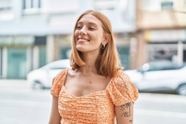 Genç kızıl saçlı kadın gülümsüyor. Kendinden emin bir şekilde caddenin kenarına bakıyor.