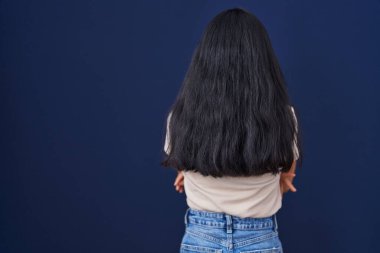 Genç İspanyol kadın mavi arka planda dikiliyor. Arkaya bakıyor ve kollarını kavuşturuyor. 