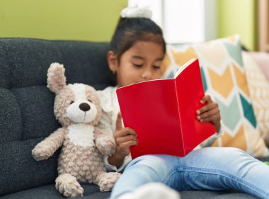 Adorable hispanic girl reading book sitting on sofa at home