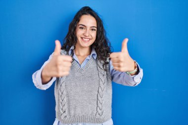 Genç esmer kadın mavi arka planda durup olumlu bir jest yapmayı onaylıyor, gülümsüyor ve başarı için mutlu. kazanan jest. 