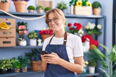 Young blonde girl florist smiling confident using smartphone at florist