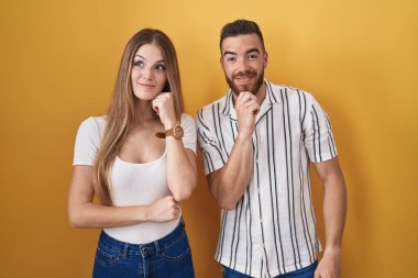 Young couple standing over yellow background with hand on chin thinking about question, pensive expression. smiling and thoughtful face. doubt concept. 