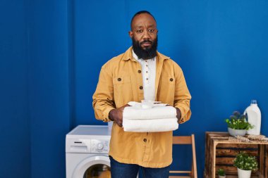 African american man holding clean towels at laundry room relaxed with serious expression on face. simple and natural looking at the camera. 