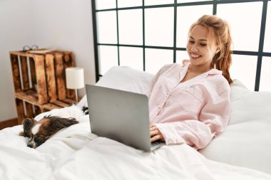Genç beyaz kadın, yatak odasında köpekle birlikte yatağında dizüstü bilgisayar kullanıyor.