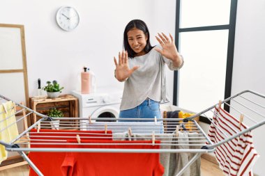 Young hispanic woman putting fresh laundry on clothesline afraid and terrified with fear expression stop gesture with hands, shouting in shock. panic concept. 