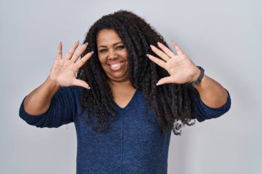 Plus size hispanic woman standing over white background showing and pointing up with fingers number ten while smiling confident and happy. 