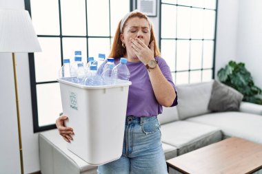 Young redhead woman holding recycling wastebasket with plastic bottles bored yawning tired covering mouth with hand. restless and sleepiness. 