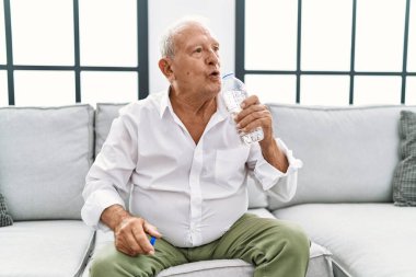 Yaşlı adam evdeki kanepede oturup su içiyor.