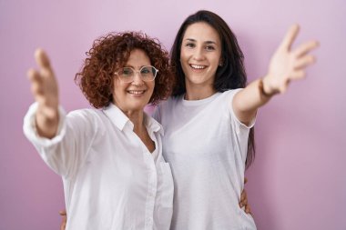 Hispanic mother and daughter wearing casual white t shirt over pink background looking at the camera smiling with open arms for hug. cheerful expression embracing happiness. 