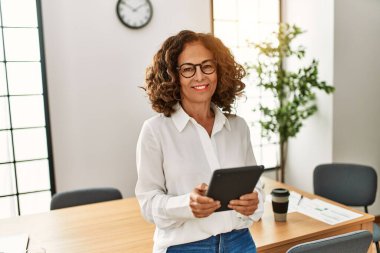 Orta yaşlı İspanyol kadın, ofiste dokunmatik ped kullanarak çalışırken kendine güvenerek gülümsüyor.