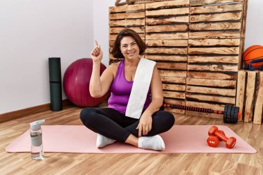Middle age hispanic woman sitting on training mat at the gym with a big smile on face, pointing with hand finger to the side looking at the camera. 