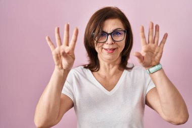 Orta yaşlı İspanyol bir kadın pembe arka planda dikilip dokuz numarayı gösterirken kendine güvenen ve mutlu bir şekilde gülümsüyor.. 