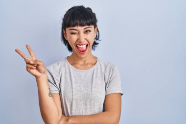 Young hispanic woman wearing casual t shirt over blue background smiling with happy face winking at the camera doing victory sign. number two. 