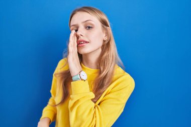 Young caucasian woman standing over blue background hand on mouth telling secret rumor, whispering malicious talk conversation  clipart