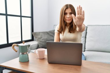 Young brunette woman using laptop at home drinking a cup of coffee doing stop sing with palm of the hand. warning expression with negative and serious gesture on the face. 