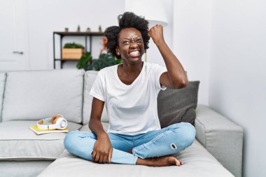 African young woman sitting on the sofa at home angry and mad raising fist frustrated and furious while shouting with anger. rage and aggressive concept. 