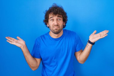 Mavi arka planda dikilen Latin kökenli genç adam elleri ve kolları havada, kafası karışmış bir ifade takınıyor. şüphe kavramı. 