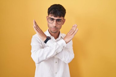 Latin asıllı genç adam sarı arka planda duruyor. Reddedilme ifadesi kollarını kavuşturuyor. Negatif işaret veriyor. 