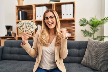 Young blonde therapist woman working at therapy office holding money screaming proud, celebrating victory and success very excited with raised arms 
