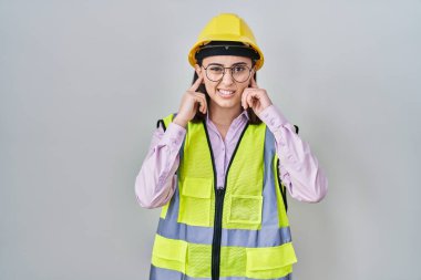 Hispanic girl wearing builder uniform and hardhat covering ears with fingers with annoyed expression for the noise of loud music. deaf concept. 