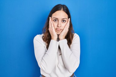 Mavi arka planda dikilen genç İspanyol kadın yorgun eller yüzünü, depresyonu ve üzüntüyü örten, sorun yüzünden üzgün ve tedirgin. 