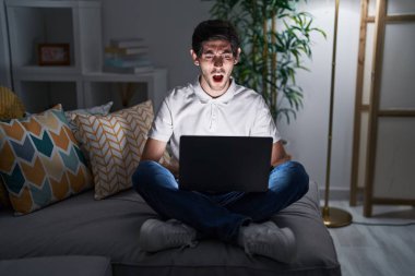Young hispanic man using laptop at home at night angry and mad screaming frustrated and furious, shouting with anger. rage and aggressive concept. 