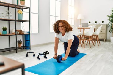 Orta yaşlı İspanyol kadın gülümsüyor. Karın kaslarını çalıştırırken evde tekerlekli sandalye kullanıyor.