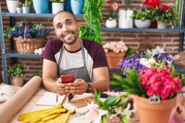 Hispanic man with beard working at florist shop with smartphone looking positive and happy standing and smiling with a confident smile showing teeth 